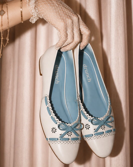 Pair of white and blue shoes with decorative elements held by a gloved hand against a beige curtain background.