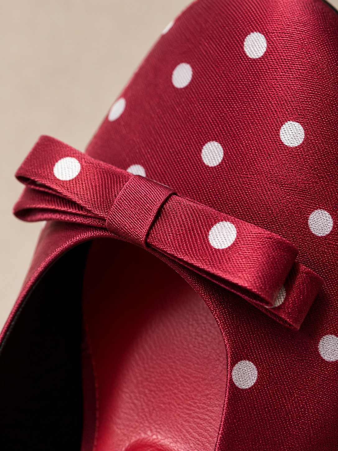 A close-up of a red satin bow with white polka dots, attached to a red fabric-covered shoe or hat on a neutral beige background.
