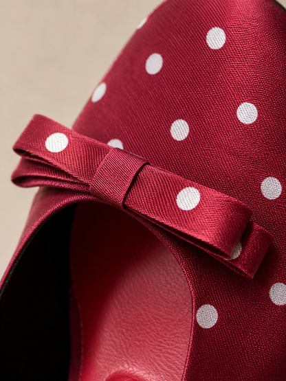A close-up of a red satin bow with white polka dots, attached to a red fabric-covered shoe or hat on a neutral beige background.