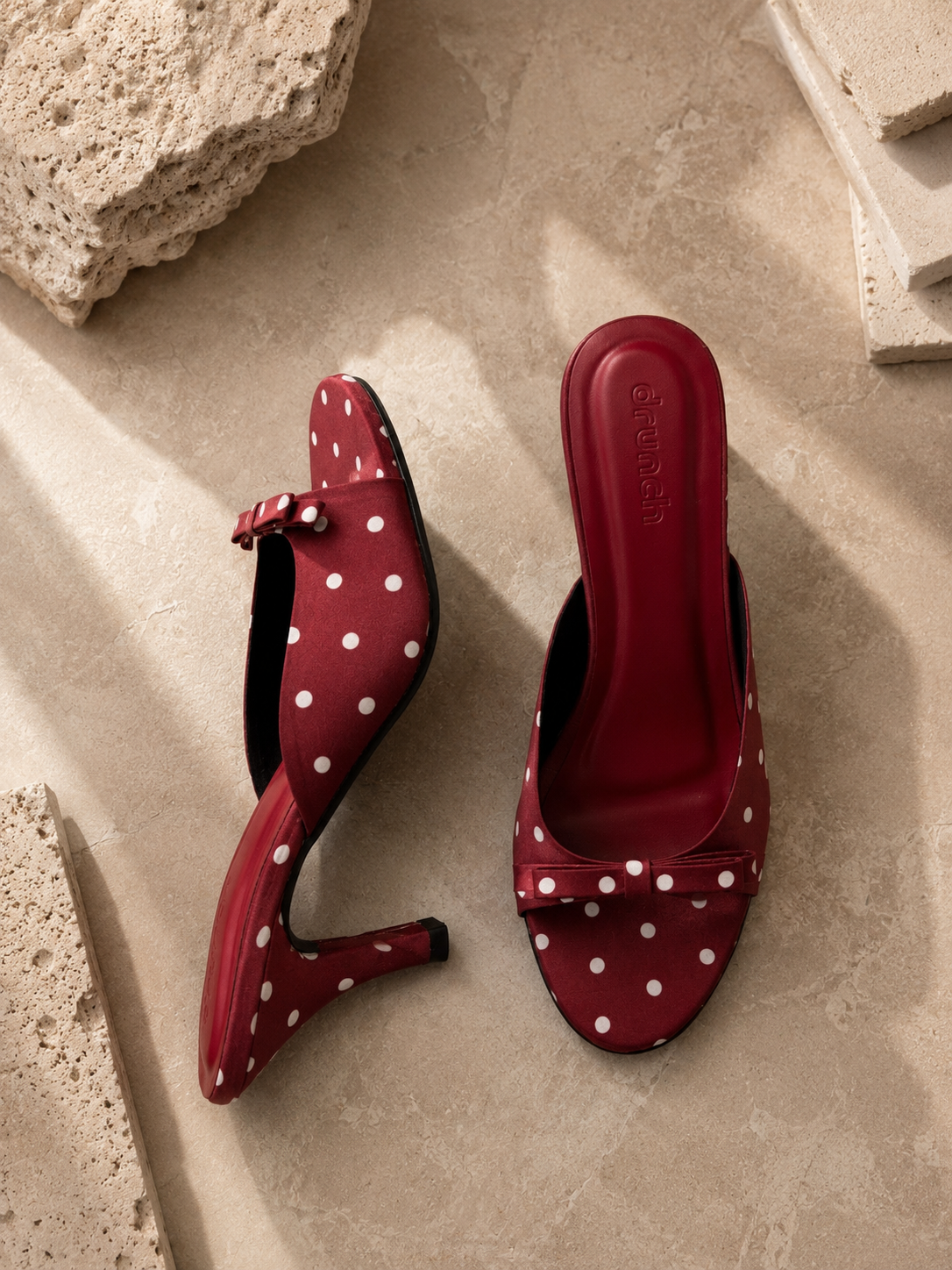 A pair of red slingback mule heels with white polka dots, displayed on a beige stone surface with decorative stone props.