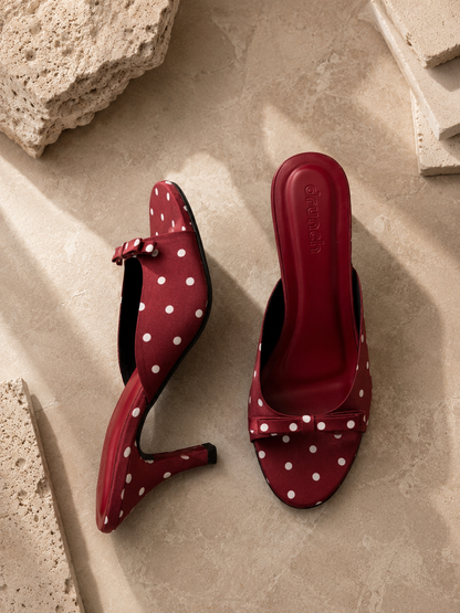 A pair of red slingback mule heels with white polka dots, displayed on a beige stone surface with decorative stone props.