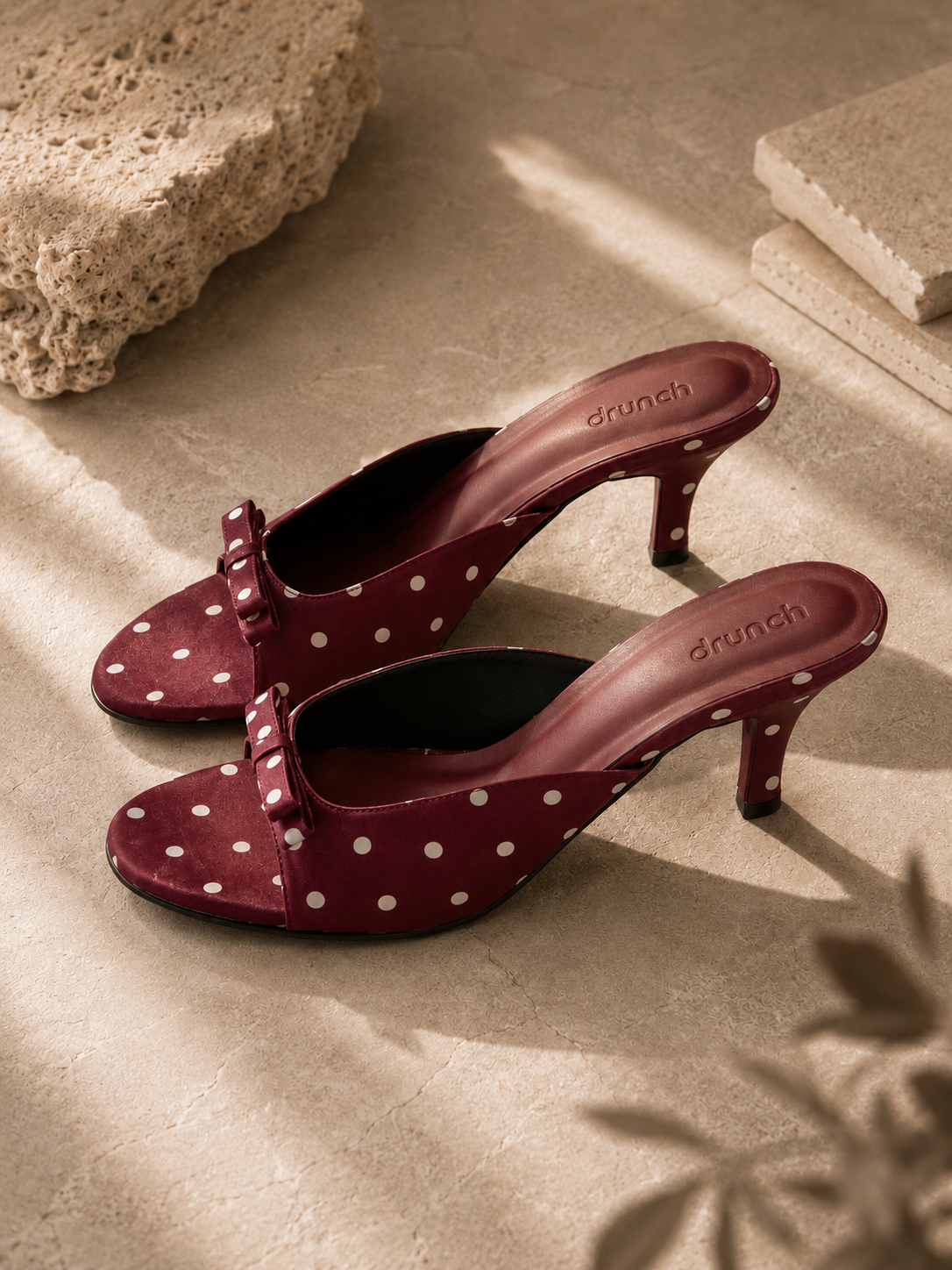 A pair of burgundy heeled mule sandals with white polka dots and bow details on the straps, displayed on a textured beige stone surface with warm sunlight and shadows.