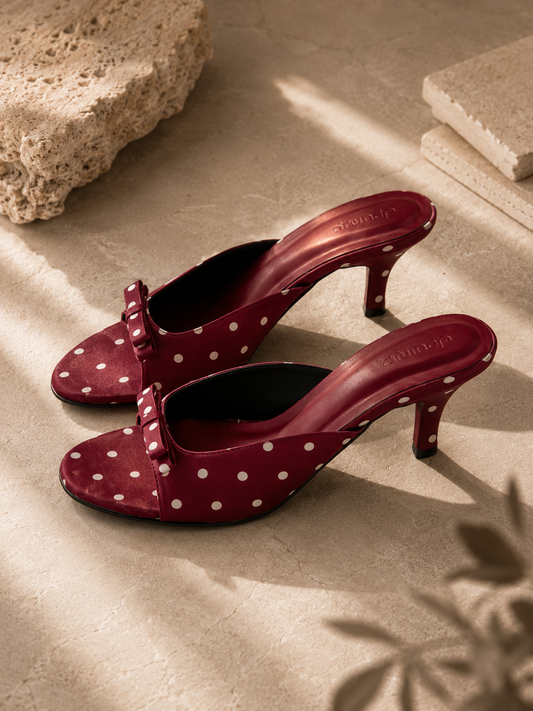 A pair of burgundy heeled mule sandals with white polka dots, styled on a beige stone surface with natural shadows.