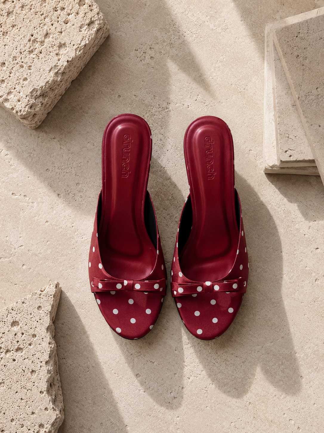 A pair of red slingback mule sandals with white polka dots and bow details, displayed on a textured beige stone surface.