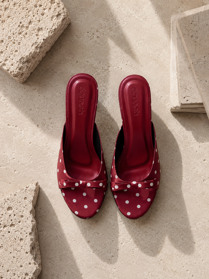 A pair of red slingback mule sandals with white polka dots and bow details, displayed on a textured beige stone surface.