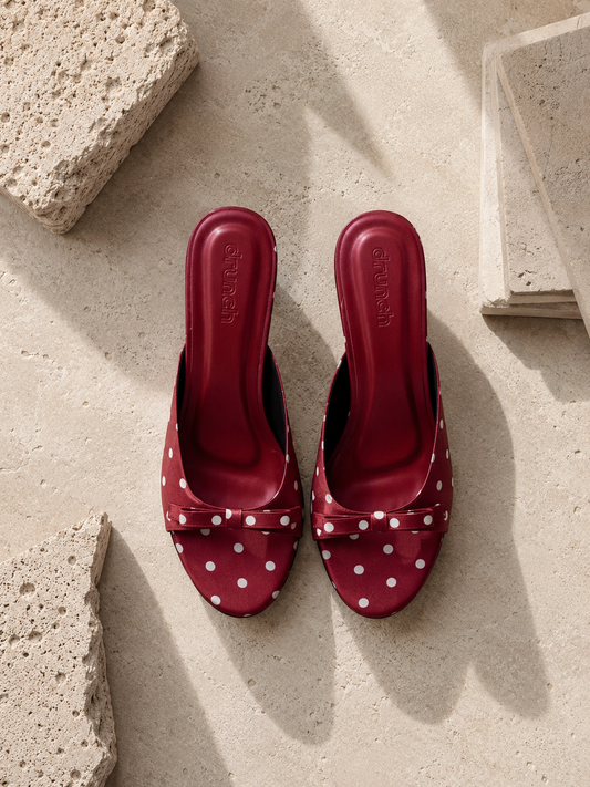 A pair of red slingback mule sandals with white polka dots and bow details, displayed on a textured beige stone surface.